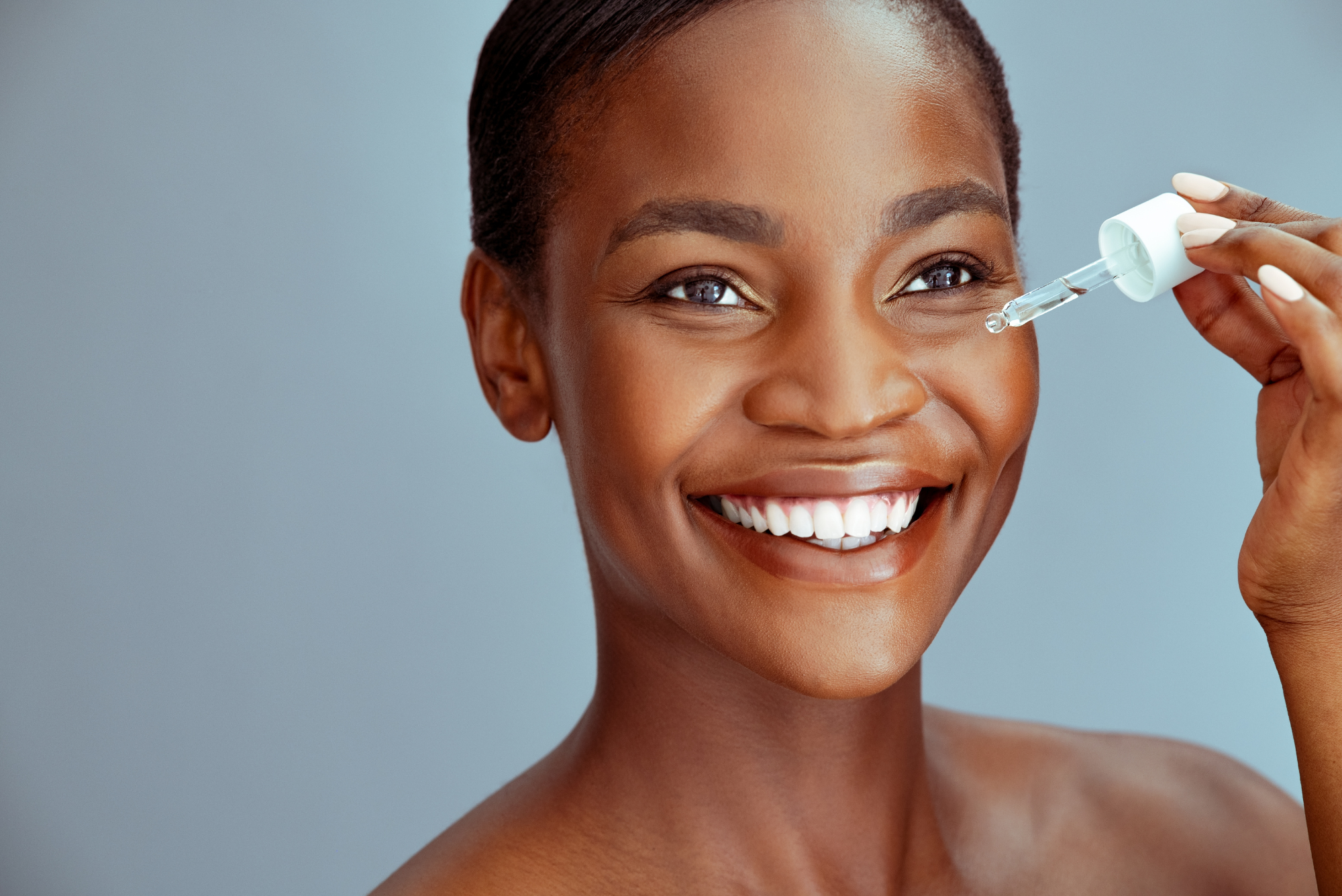 A woman smiles as she applies a deep-penetrating led peptide serum to her cheek with a dropper.