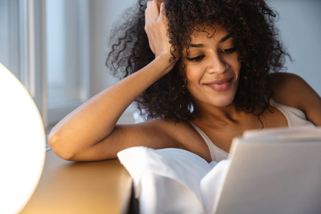 A woman with curly hair lies in her bed next to a lamp, reading a book about natural treatments for genital herpes.